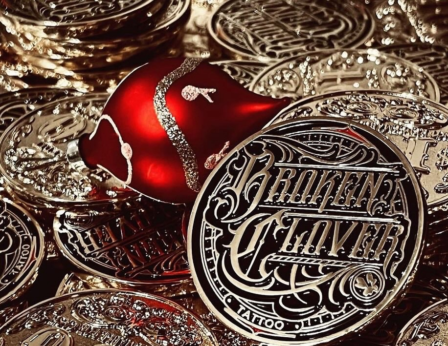 Gold-toned real Broken Clover Tattoo Gift Coins with an ornate embossed logo and red holiday ornament.