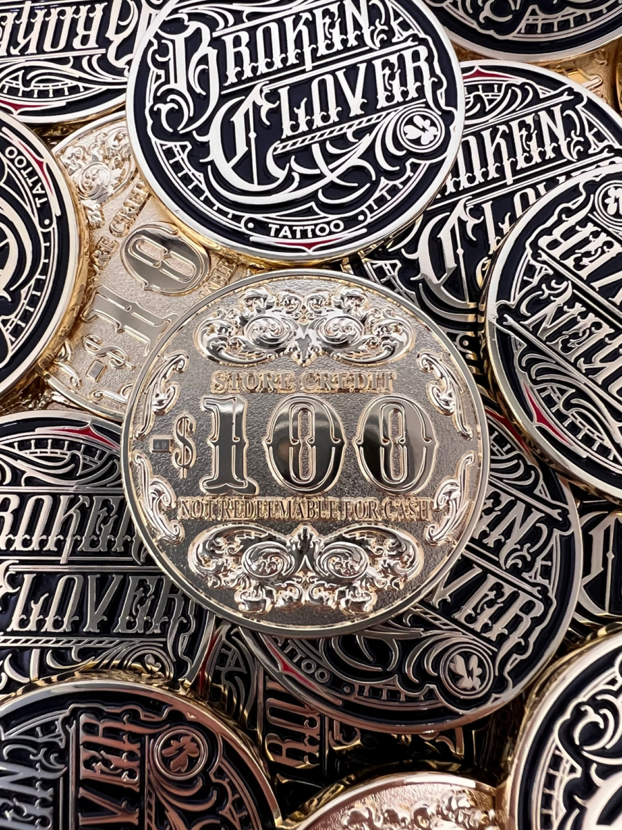 Photograph of physical Broken Clover Tattoo gift coins piled together, showing the ornate shop logo and the reverse side marked $100 store credit.